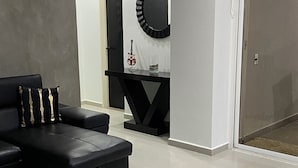 A modern living room with a black sofa, a black chair, and a black console table. There is a round mirror and a clock on the wall. The room has white walls and a tiled floor.
