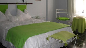 A neatly made bed with white bedding and a green throw, a small bedside table with a green cushion, a white chair, and a small table with a green tablecloth. - Noria Hotel (Villa Carlos Paz)