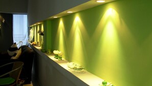 A long, narrow room with a green wall and a long counter. The counter has various items on it, including a vase with flowers, a bowl, and a green apple. There is a chair with a green cushion and a small table with a green object on it. - Noria Hotel (Villa Carlos Paz)