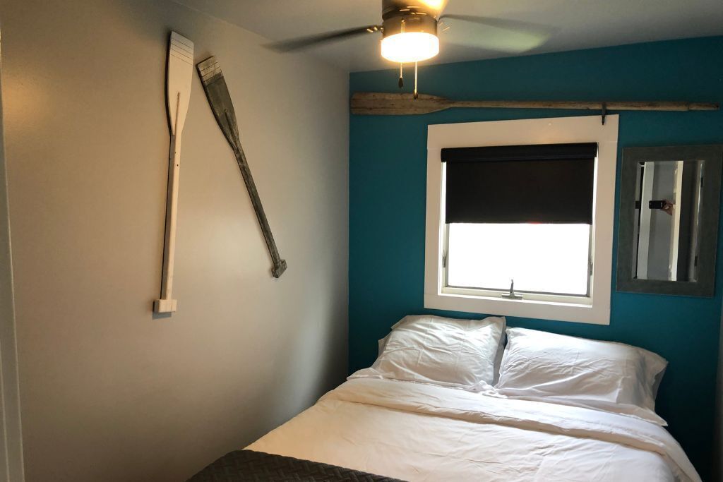 A bedroom with a bed, a window with a white blind, a ceiling fan, and a wall-mounted light fixture.