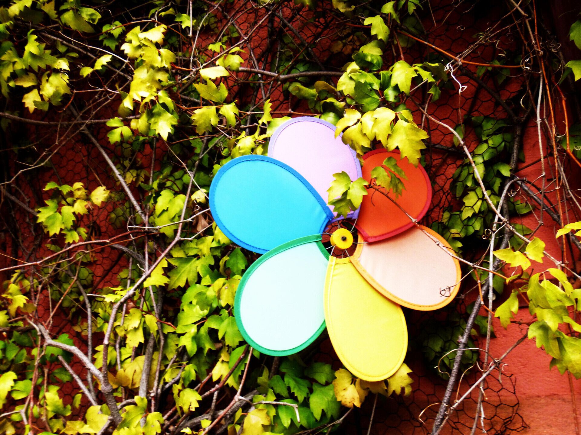A colorful windmill-like structure with multiple colored blades, surrounded by green ivy and a red brick wall.