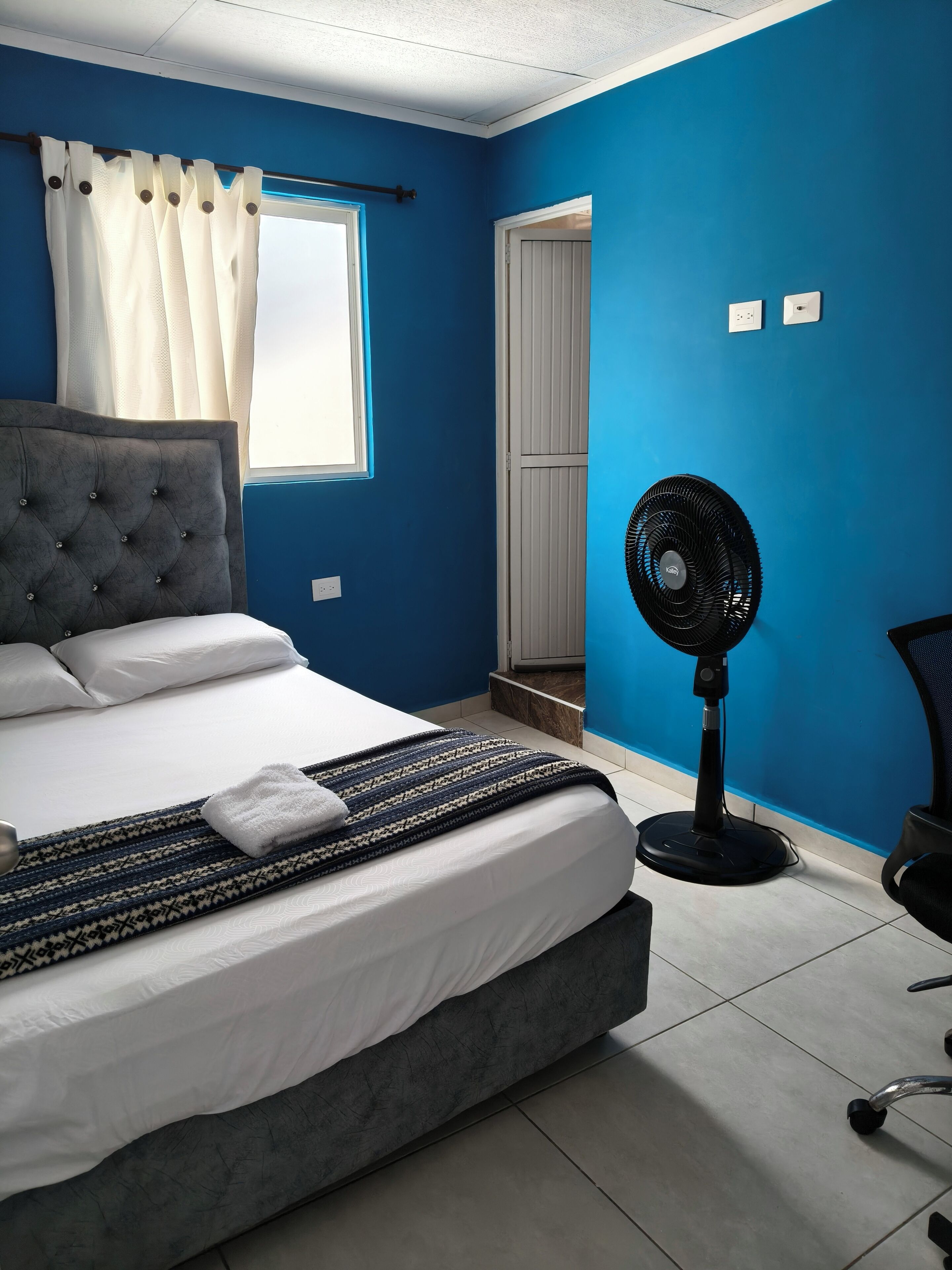 A room with a bed, a standing fan, a chair, and a door leading to another room.