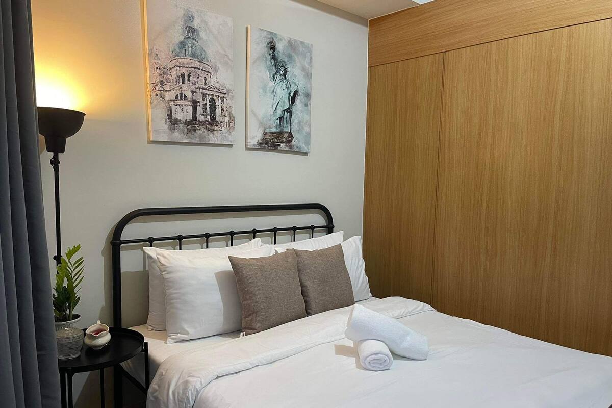 A neatly made bed with white linens and a rolled towel, a black metal headboard, and two paintings of architectural structures on the wall.