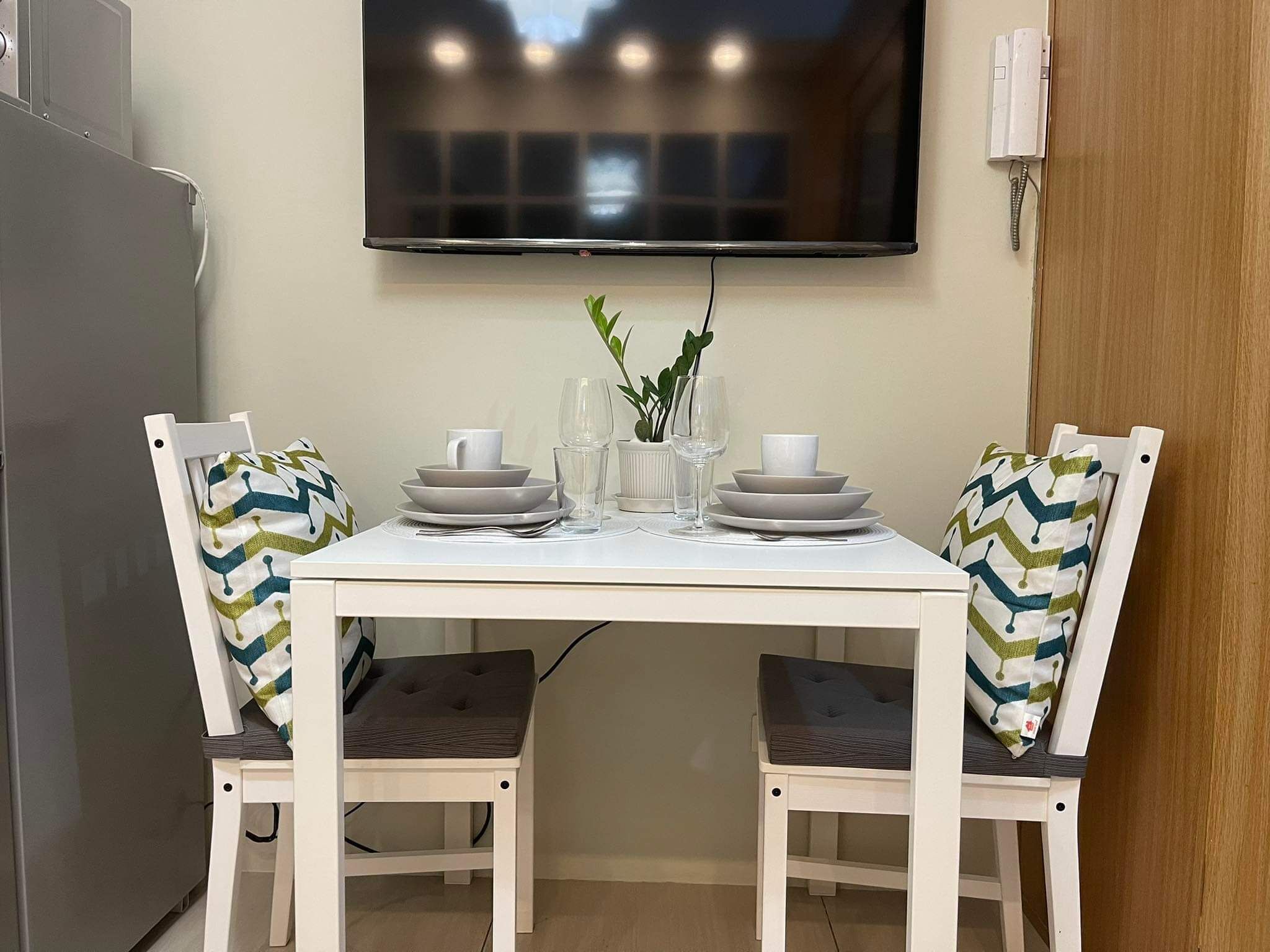 A dining area with a white table set for two, two chairs with patterned cushions, a flat-screen TV mounted on the wall, and a small potted plant on the table.