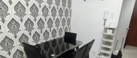 Un comedor con una mesa de vidrio y sillas negras, un papel tapiz estampado, un estante blanco y un zapatero.