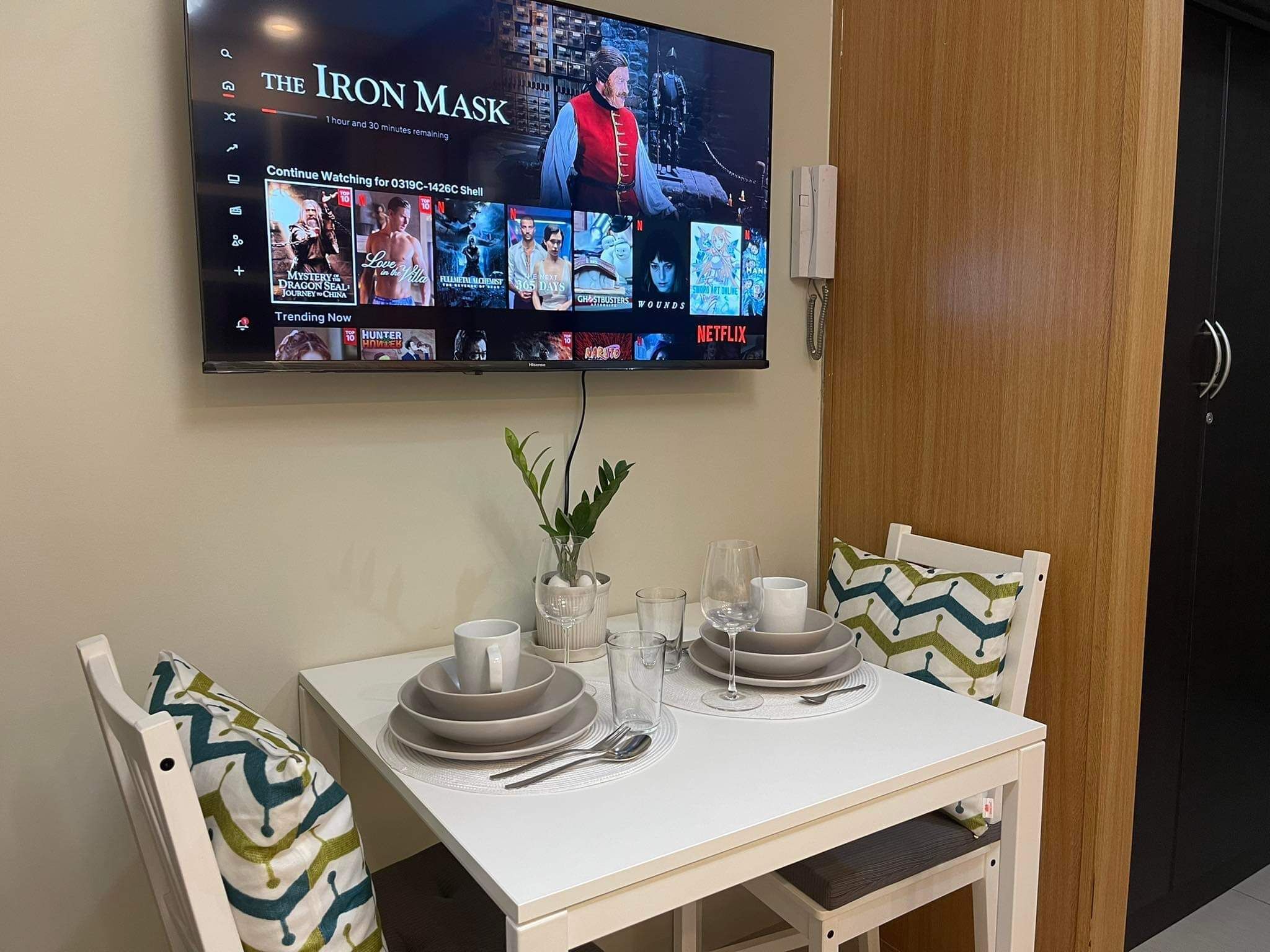A dining area with a TV mounted on the wall displaying 'The Iron Mask'.