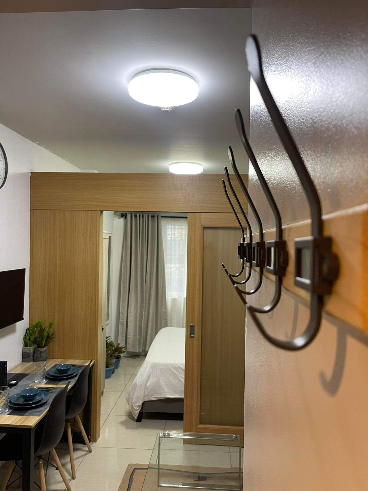 A modern hotel room with a wooden door, a dining area with a table and chairs, a bed, and a ceiling light.