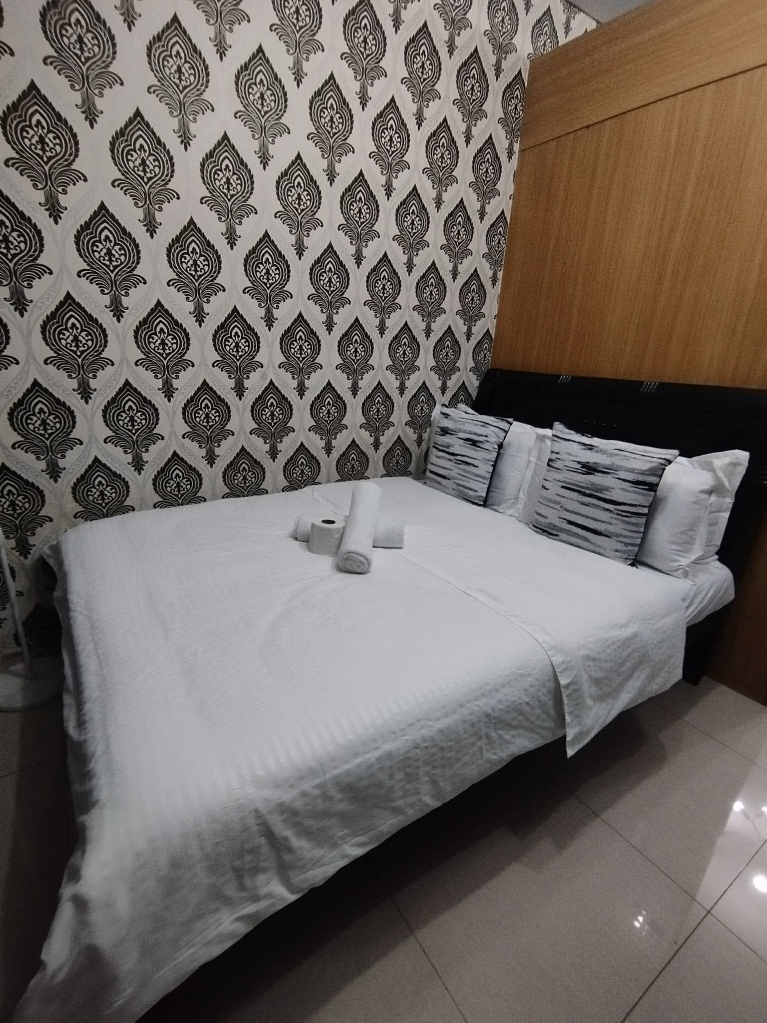 Una cama bien hecha con ropa blanca y toallas enrolladas, un papel tapiz estampado y un cabecero de madera.