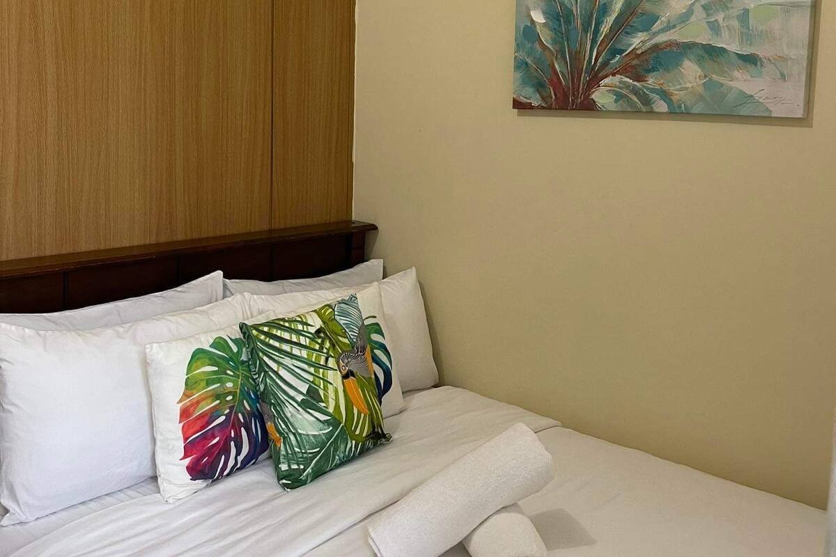 A hotel room with a bed, white linens, a wooden headboard, a tropical-themed pillow, and a painting of a plant on the wall.