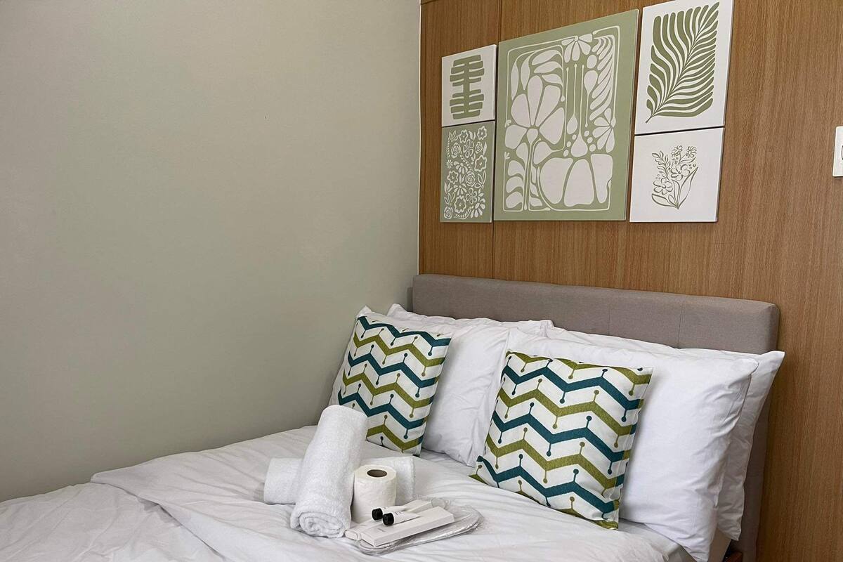 A neatly made bed with white linens, two pillows with a chevron pattern, a rolled towel, and a roll of toilet paper.