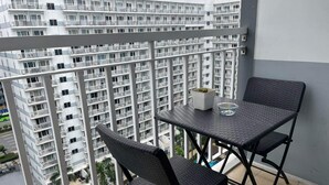 Een balkon met twee zwarte rieten stoelen en een kleine tafel, met uitzicht op een zwembad en een hoog gebouw met balkons.