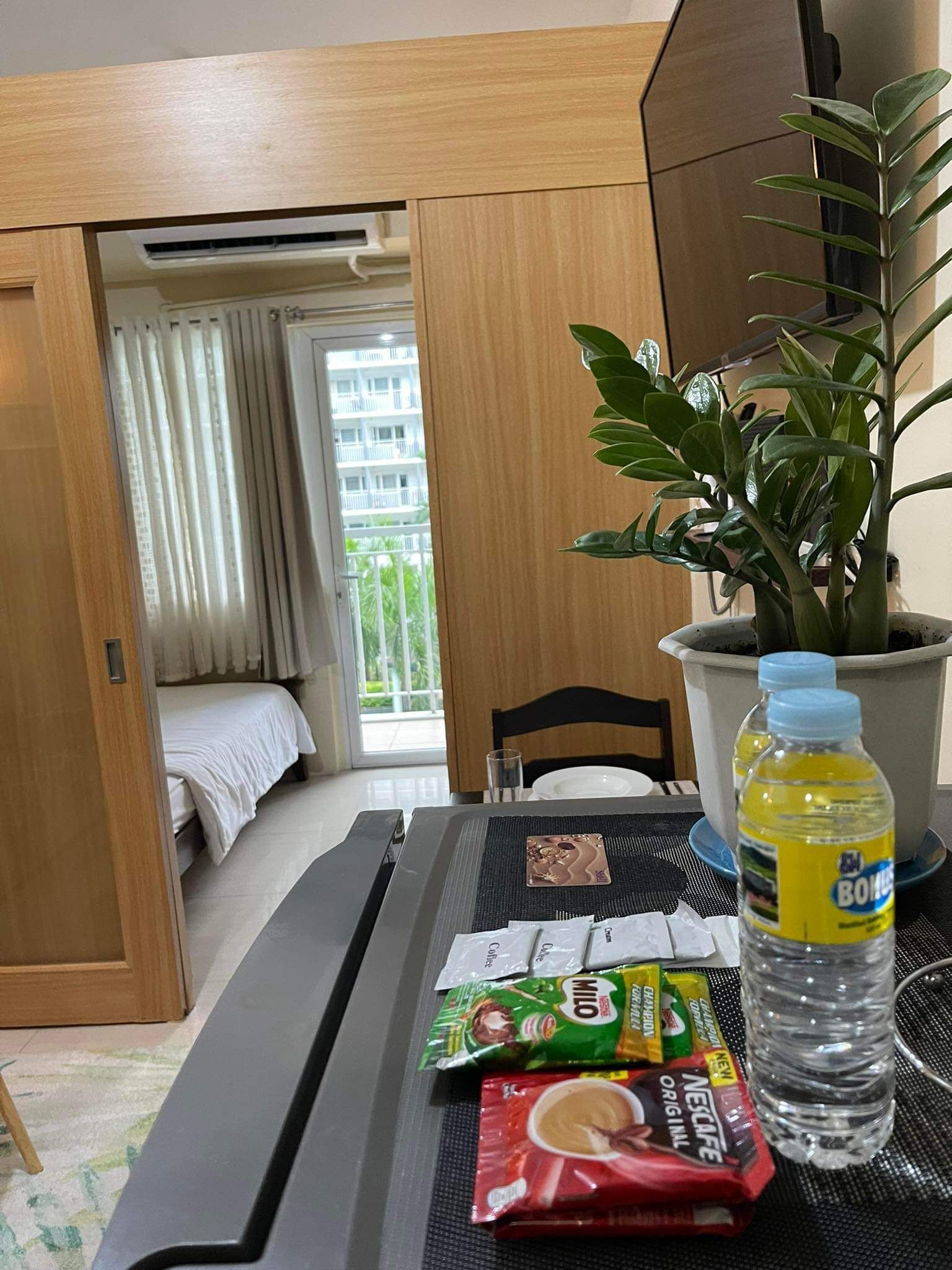 A hotel room with a bed, a dining table with a placemat, a potted plant, a bottle of water, and a packet of instant coffee.