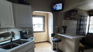 A compact kitchen with white cabinets, a built-in sink, and a small bar area with a stool. A television is mounted on the wall above the bar.