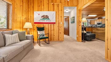 Een knus ingerichte kamer met houten details, een vlag aan de muur, een schommelstoel, een bank en een keukenhoek.