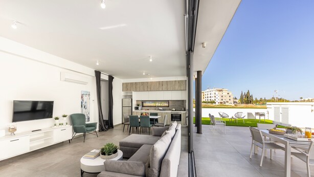 A modern living room with a flat-screen TV, grey sofas, and a dining area with white furniture.