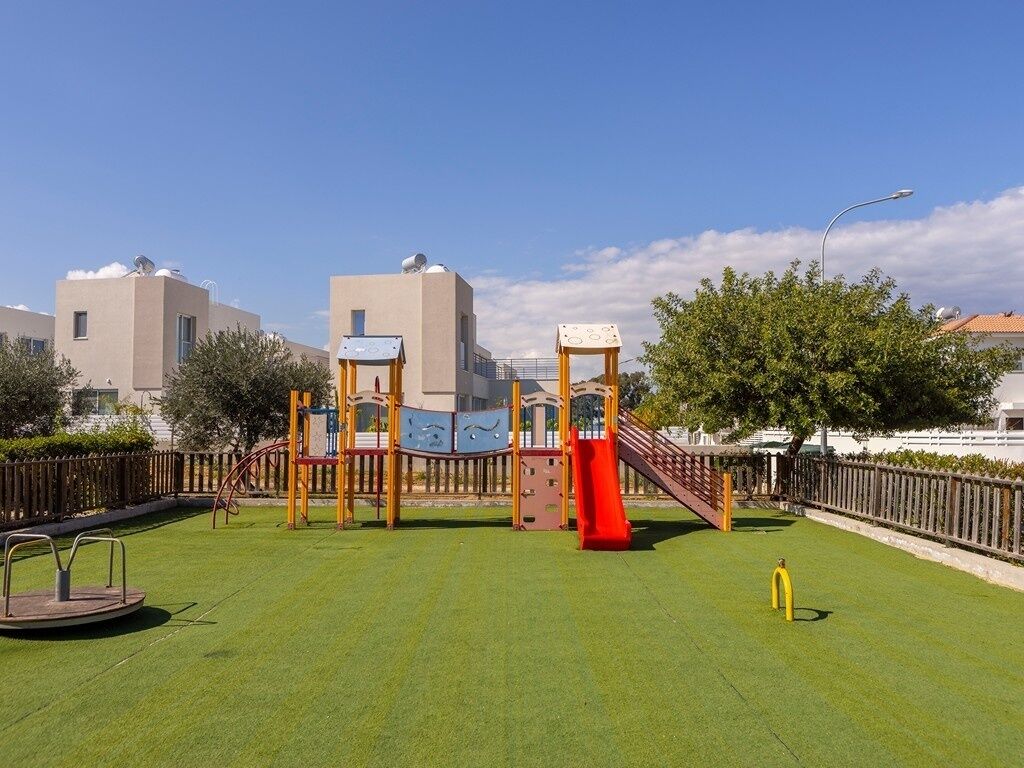 Une aire de jeux avec un toboggan et des structures d’escalade, entourée d’une clôture et de verdure.