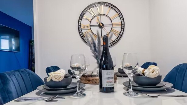 Une table à manger est dressée avec des verres à vin, une bouteille de vin et des bols remplis de nourriture. Un grand cadran d’horloge est accroché au mur, derrière la table.