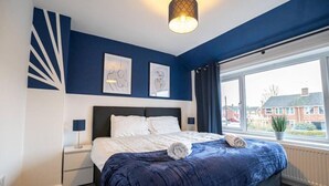 Une chambre à coucher avec un lit, une table de chevet, une lampe, une fenêtre avec des rideaux et des œuvres d’art accrochées au mur.