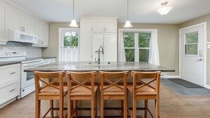 A kitchen with white cabinets, a large island with bar stools, pendant lighting, and a view of the outdoors through a window.