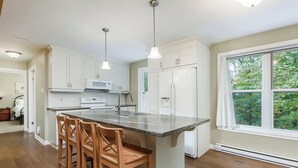 A modern kitchen with a central island, white cabinetry, stainless steel appliances, and a large window offering a view of greenery.