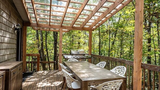 A wooden deck with a table and chairs, a grill, and a pergola with glass panels.