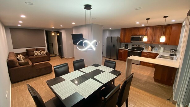 A modern kitchen with wooden cabinets, a dining area with a table and chairs, and a living room with a sofa and a TV.