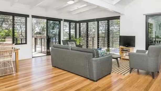 A spacious living room with a large grey sofa, a wooden coffee table, and a TV stand. The room has large windows and a ceiling fan.