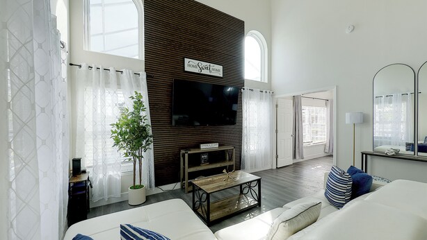 A modern living room with a large flat-screen TV mounted on a wooden wall, a white sofa, a coffee table, and a potted plant. - Themed Family Retreat w/ Pool, Game Room – Minutes to Disney! (Kissimmee)