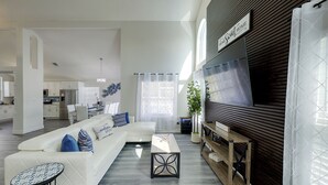 A modern living room with a white sofa, a coffee table, and a TV on a wooden wall. - Themed Family Retreat w/ Pool, Game Room – Minutes to Disney! (Kissimmee)