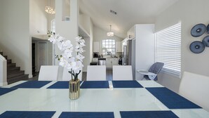 A dining area with a glass table, blue placemats, white chairs, and a vase of white flowers. - Themed Family Retreat w/ Pool, Game Room – Minutes to Disney! (Kissimmee)
