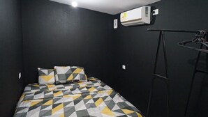 Ein kleiner, dunkel eingerichteter Raum mit einem Bett, das eine geometrische Tagesdecke hat, einer Wand-Klimaanlage und einer Metallleiter an der Wand.