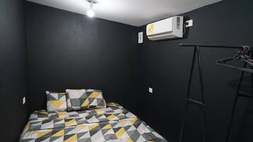 Una habitación pequeña y oscura con una cama que tiene una colcha de estampado geométrico, un aire acondicionado de pared y una escalera metálica apoyada contra la pared.