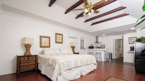 Une chambre avec un grand lit, des tables de chevet en bois, un ventilateur de plafond et, au fond, un coin cuisine.