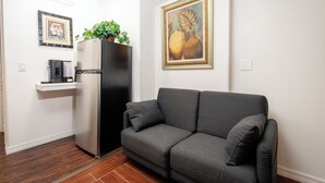 Un salon avec un canapé gris, un réfrigérateur noir, une machine à café et un tableau encadré accroché au mur.