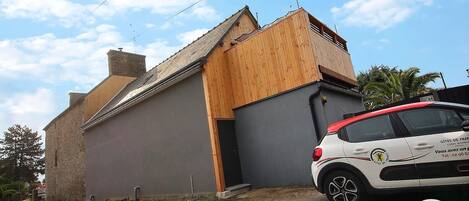 Un bâtiment à deux étages, doté d’une façade grise et d’une extension en bois. Une voiture blanche, portant l’inscription « Gîtes de France », est garée devant.