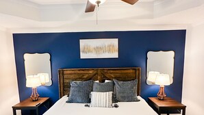 A bedroom with a large bed, two nightstands, a ceiling fan, and a painting on the wall.