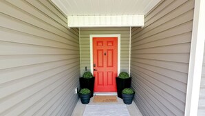 A covered walkway leading to a red door with two potted plants on either side.