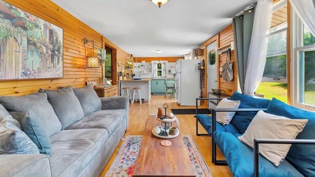 A cozy living room with a grey sofa, a blue sofa, and a wooden coffee table. There is a kitchen in the background with a fridge and a sink. - Point Prim Cottage: Beach Access and Family Fun (Belfast)