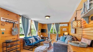 A cozy cabin-style room with wooden walls, a sofa, a chair, a small table, and a hanging light fixture. - Point Prim Cottage: Beach Access and Family Fun (Belfast)