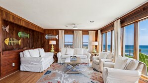 A spacious room with a wooden wall, white furniture, a large window with a view of the ocean, and a colorful rug. - Beachfront Duck retreat with 4 king bedrooms, 3 baths, and private beach walkway. (Duck)