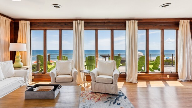 A spacious living room with a large window overlooking the ocean, a wooden ceiling, and a cozy seating area with a coffee table and chairs. - Beachfront Duck retreat with 4 king bedrooms, 3 baths, and private beach walkway. (Duck)