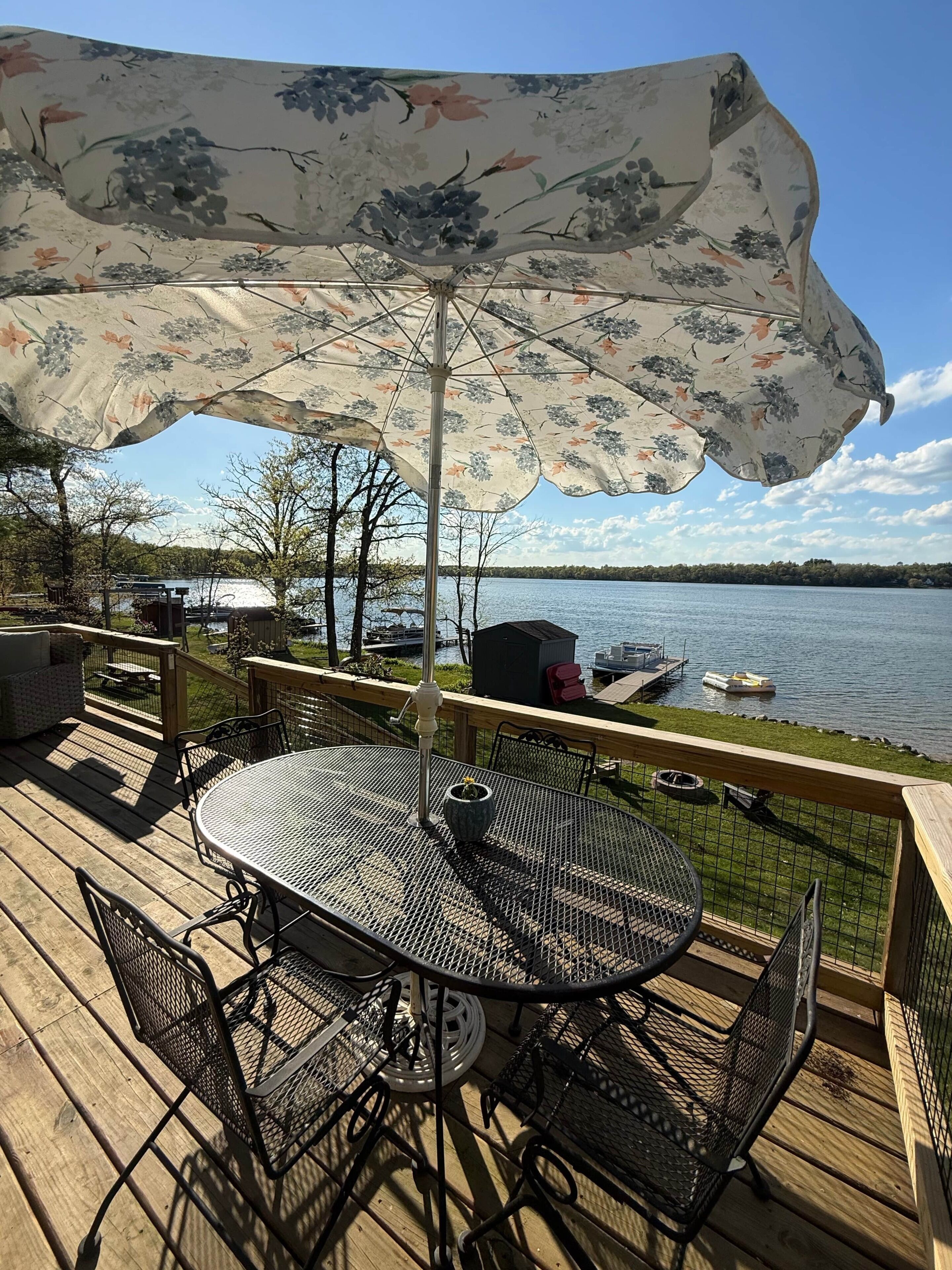 A deck with a table and chairs under a floral umbrella, overlooking a lake with boats.