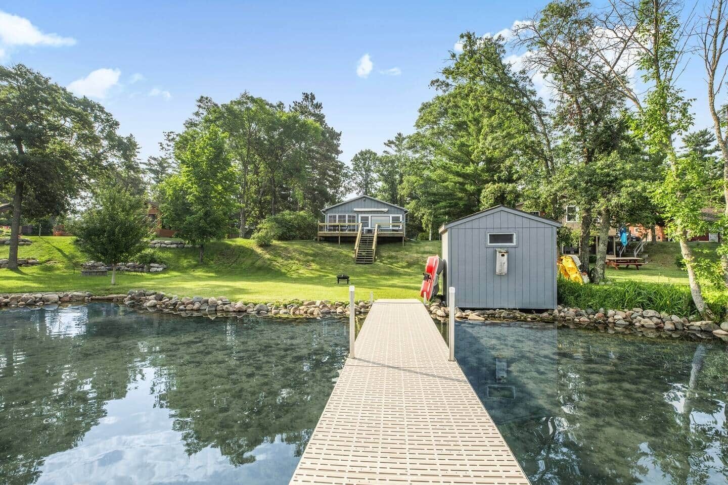 Eine Holzsteg, der zu einem Haus mit einem kleinen Schuppen führt, umgeben von einem Teich und viel Grün.