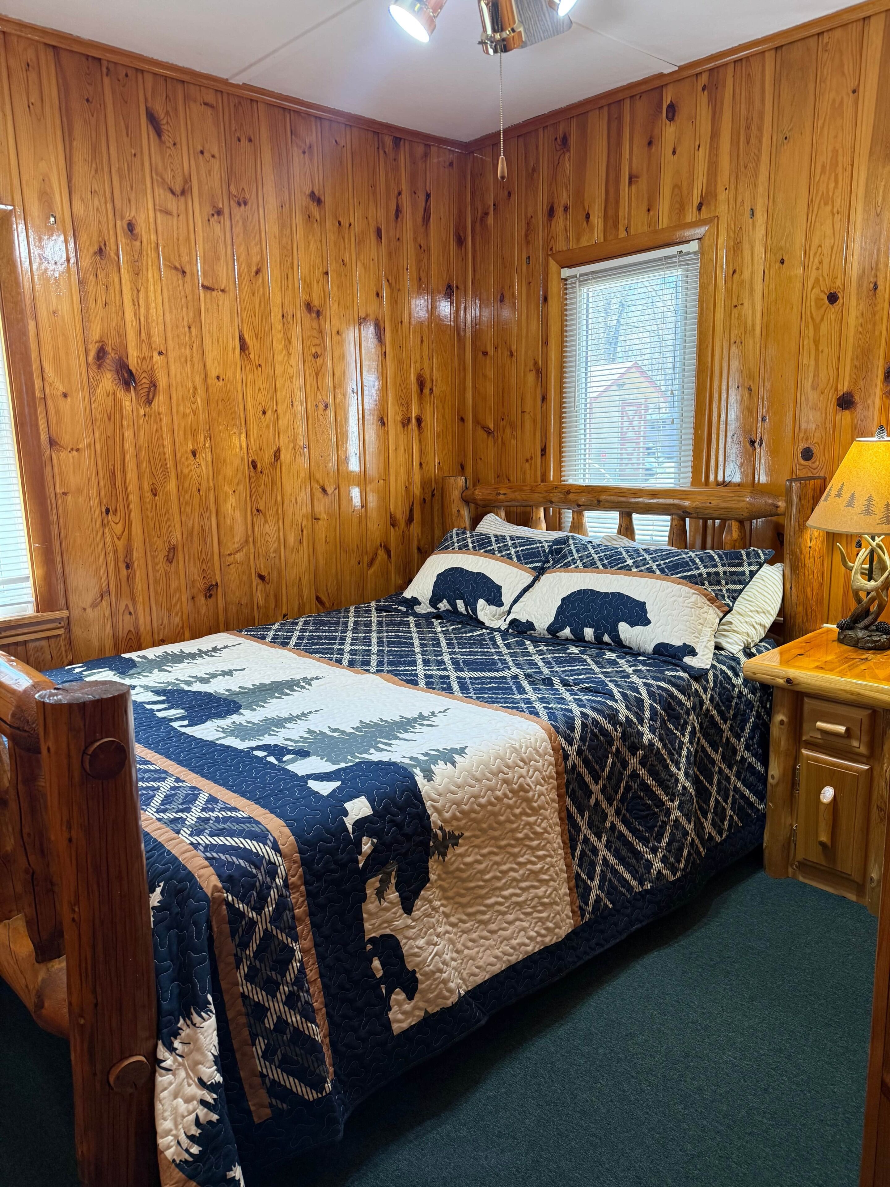 A wooden-paneled room with a bed, a nightstand, and a window with blinds.