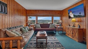 A cozy cabin-style room with a wooden interior, a large window offering a view of the outdoors, a sofa, a coffee table, and a cabinet with decorative items. - The Cozy Cub (Crosslake)