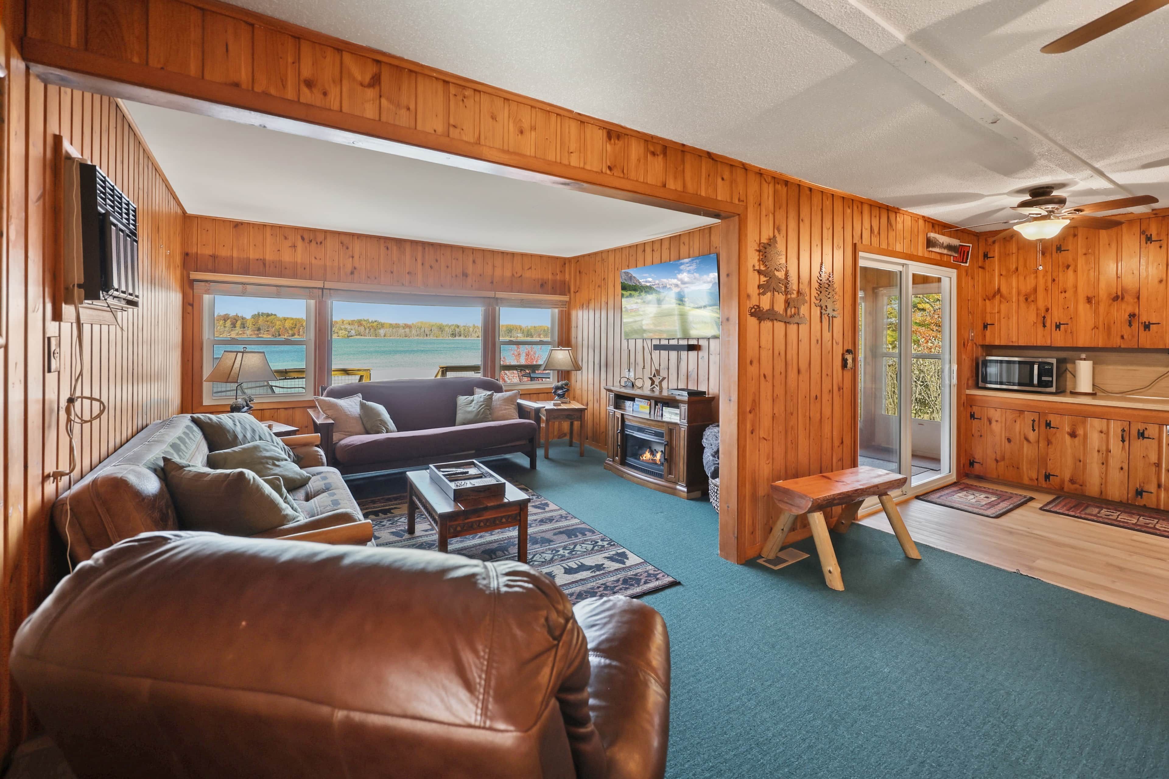 A cozy cabin interior with a living room, a fireplace, a dining area, and a view of a lake.