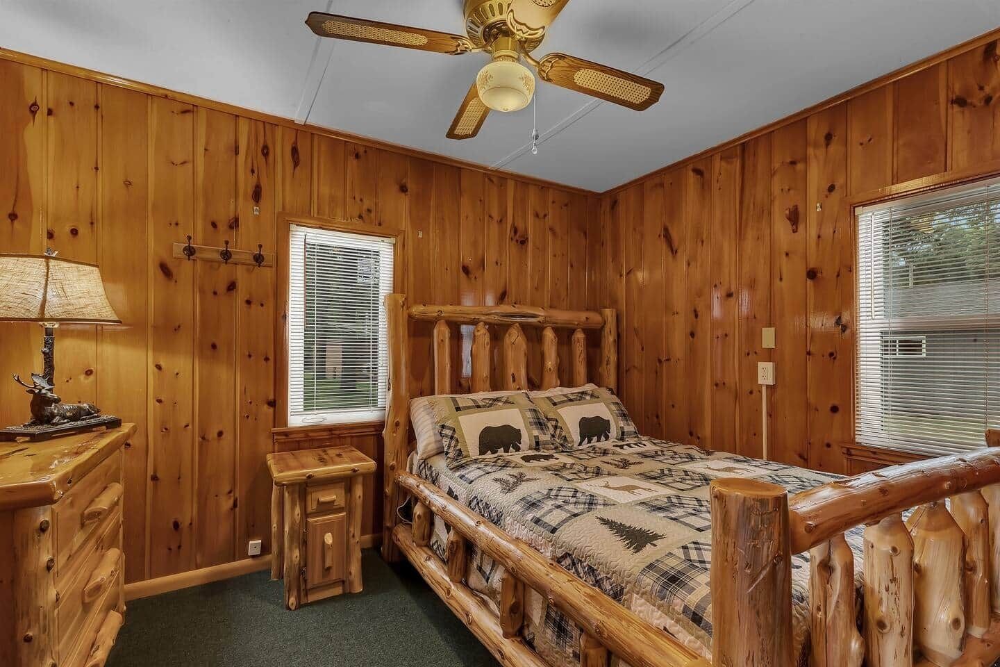 Dormitorio de cabaña de madera con cama, mesita de noche, lámpara, ventilador de techo y dos ventanas con persianas.