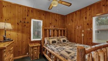 Dormitorio de cabaña de madera con cama, mesita de noche, lámpara, ventilador de techo y dos ventanas con persianas.