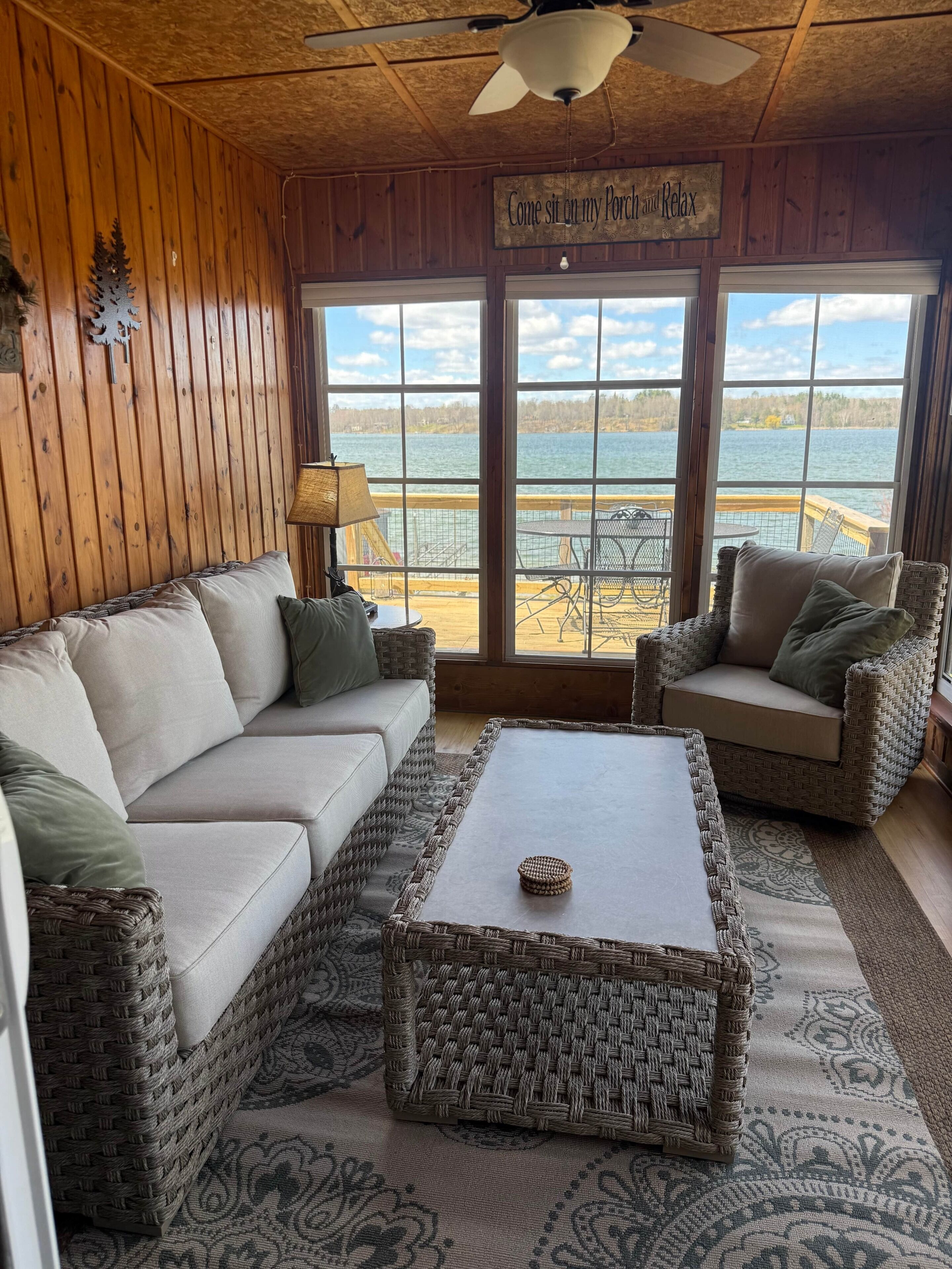 A cozy room with a wooden ceiling, a ceiling fan, a large window with a view of a lake, a wicker sofa, a wicker armchair, and a small table with a lamp.