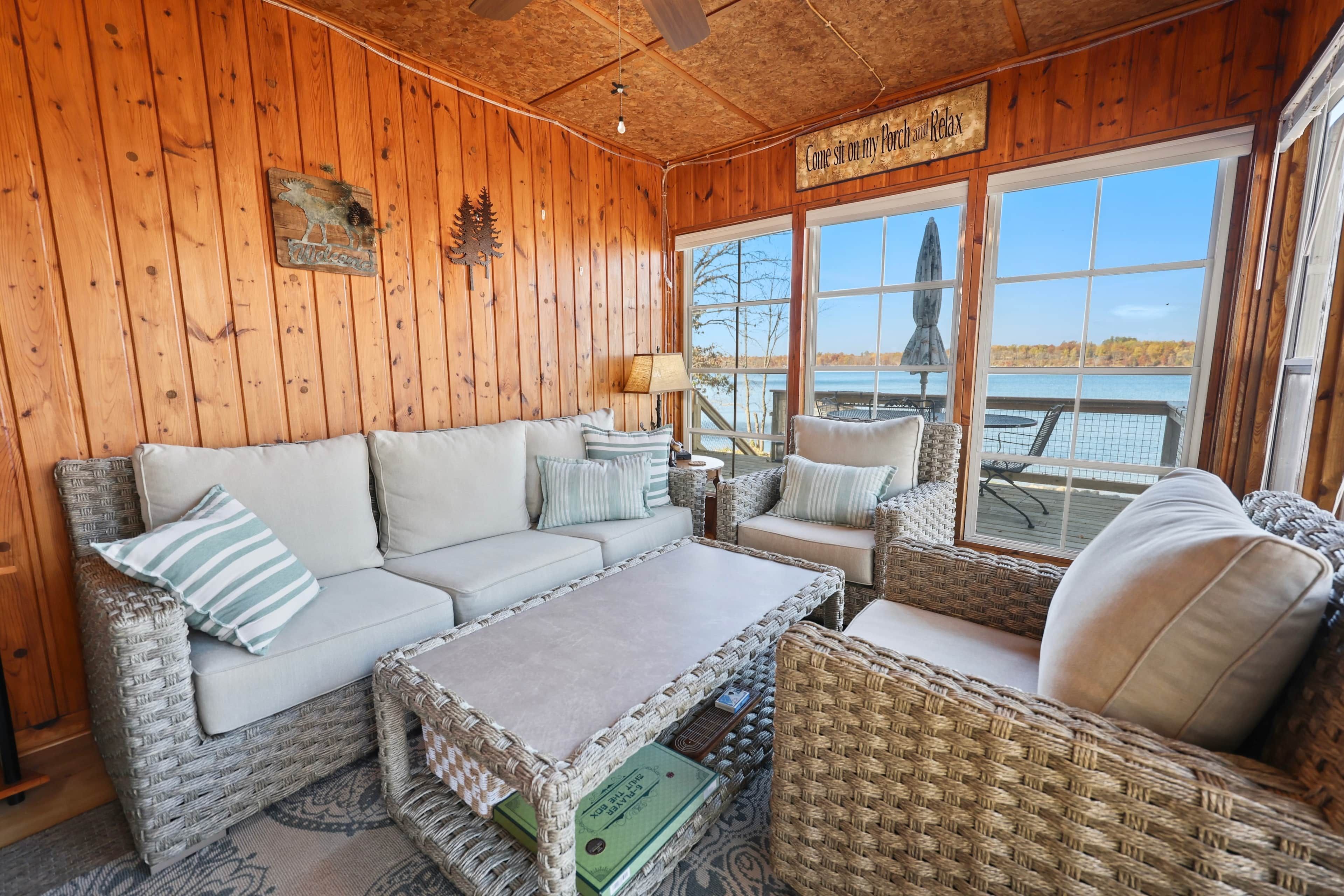 A cozy cabin interior with a sofa, chairs, a table, and a view of a lake through large windows.